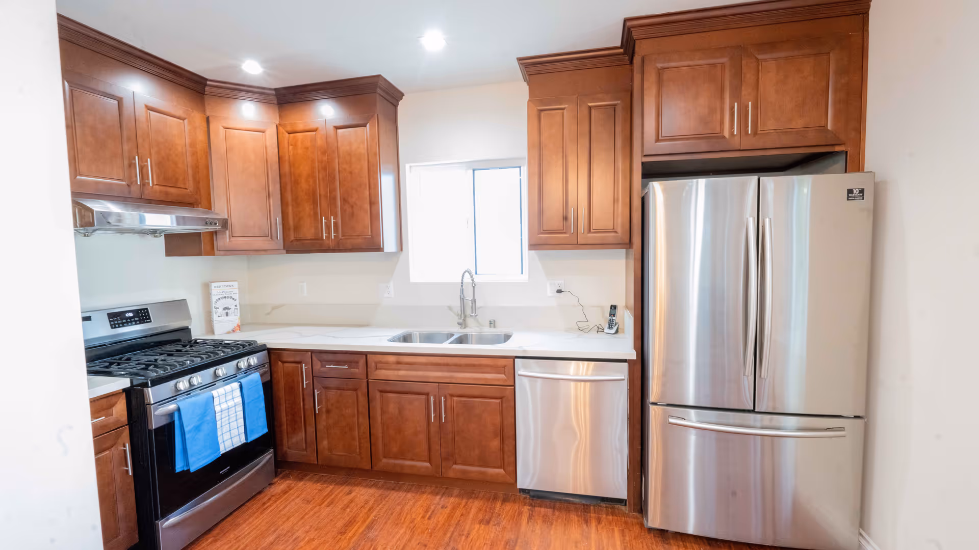 A modern kitchen with wooden cabinets, a stainless steel refrigerator, a dishwasher, a gas stove with two blue towels hanging on the handle, and a double sink under a window. The floor is wooden and the walls are light-colored.