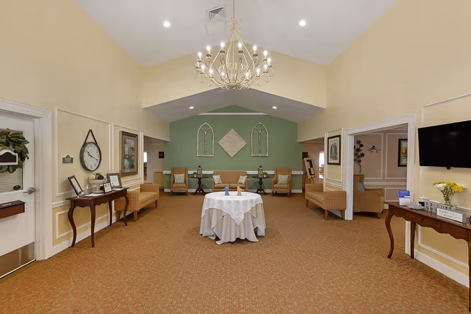 Spacious senior living common lounge with upholstered chairs, side tables, a round table covered with a white cloth under a chandelier and a green accent wall with decor.