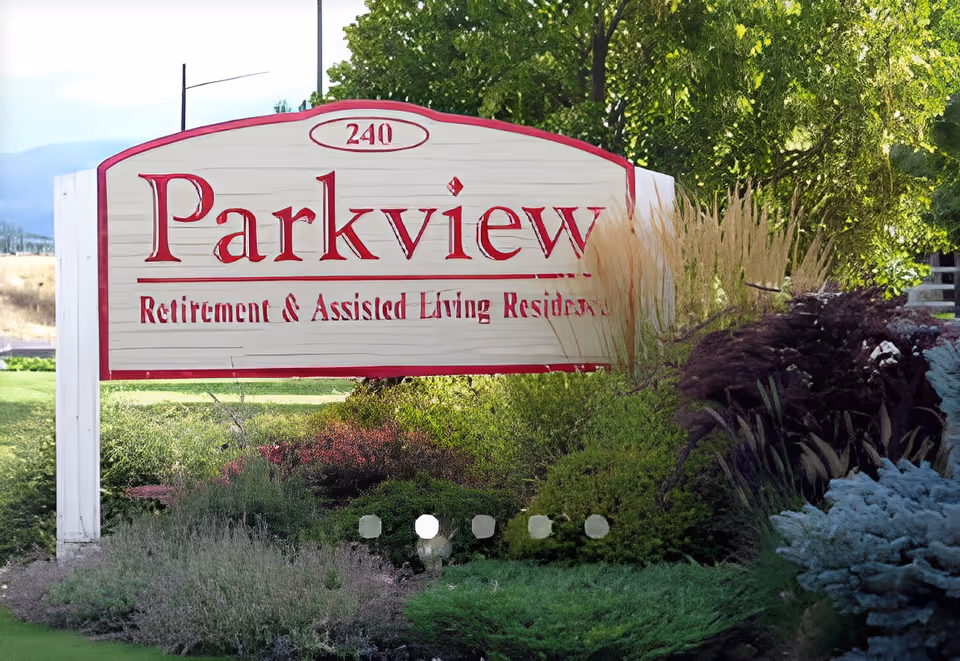 A large wooden sign with red lettering that reads 'Parkview Retirement & Assisted Living Residence' with the number 240 above it. The sign is surrounded by green bushes, ornamental grasses, and trees in a landscaped outdoor area.