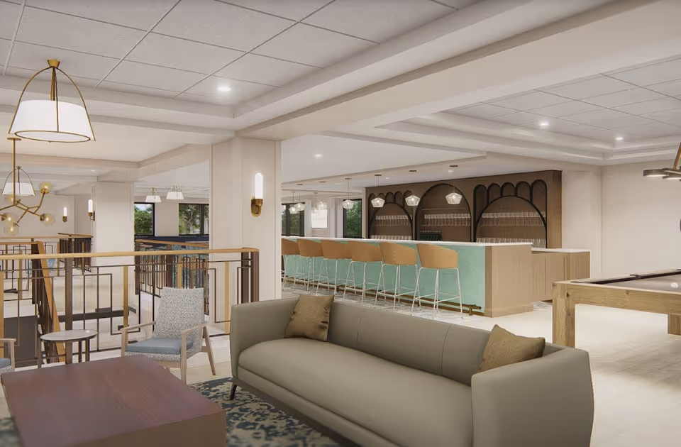A modern senior living facility common area featuring a gray sofa with brown pillows, a wooden coffee table, a single armchair, and a bar with several tan bar stools. The space is well-lit with ceiling lights and pendant lamps, and there is a pool table visible on the right side. The area has a neutral color palette with light walls and flooring, and large windows letting in natural light.