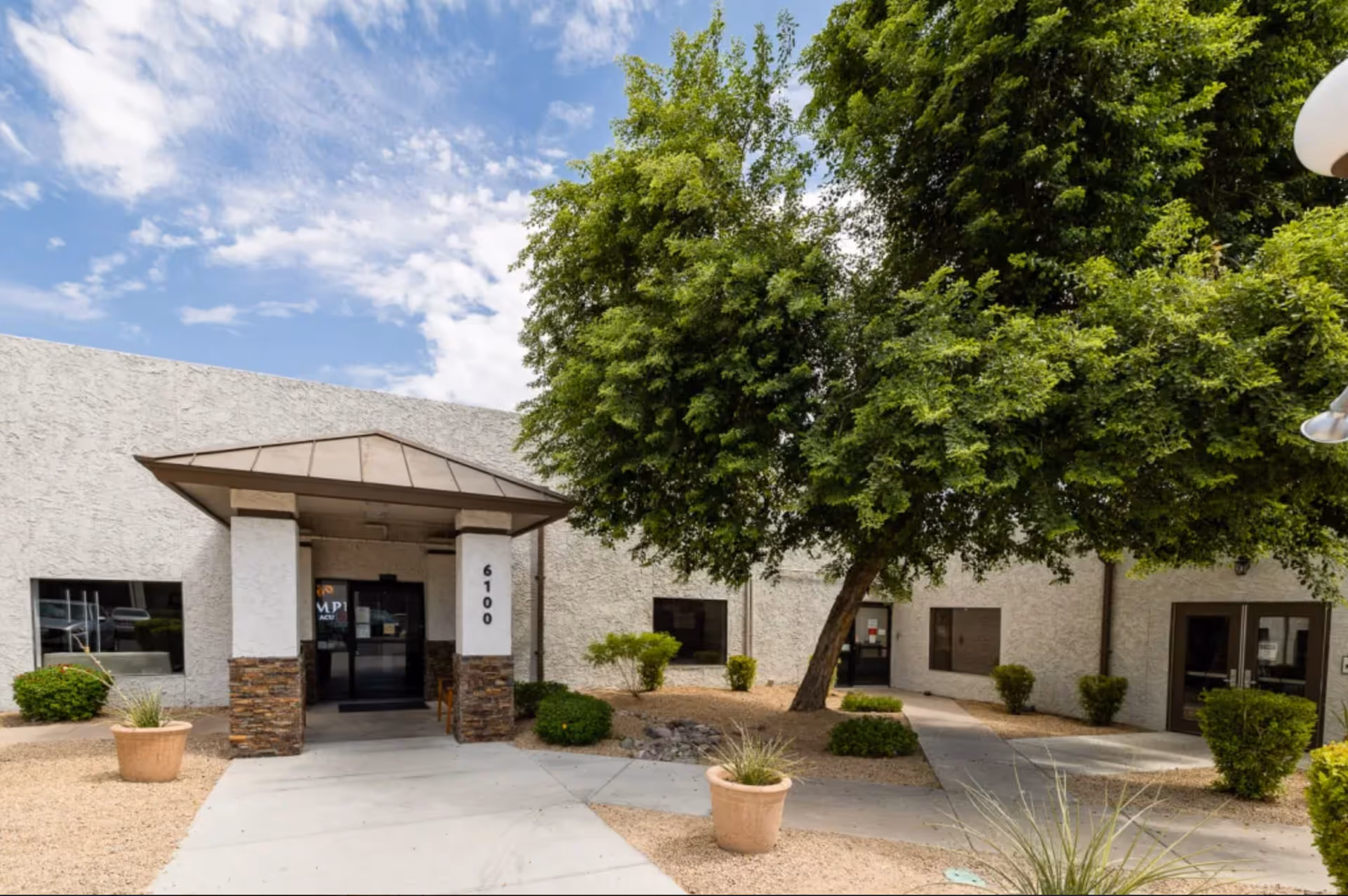 Exterior view of a single-story building with a covered entrance supported by two pillars. The building has a textured white facade with several windows and doors. There is a large green tree and some potted plants and bushes in a landscaped area with gravel and concrete walkways under a partly cloudy sky.