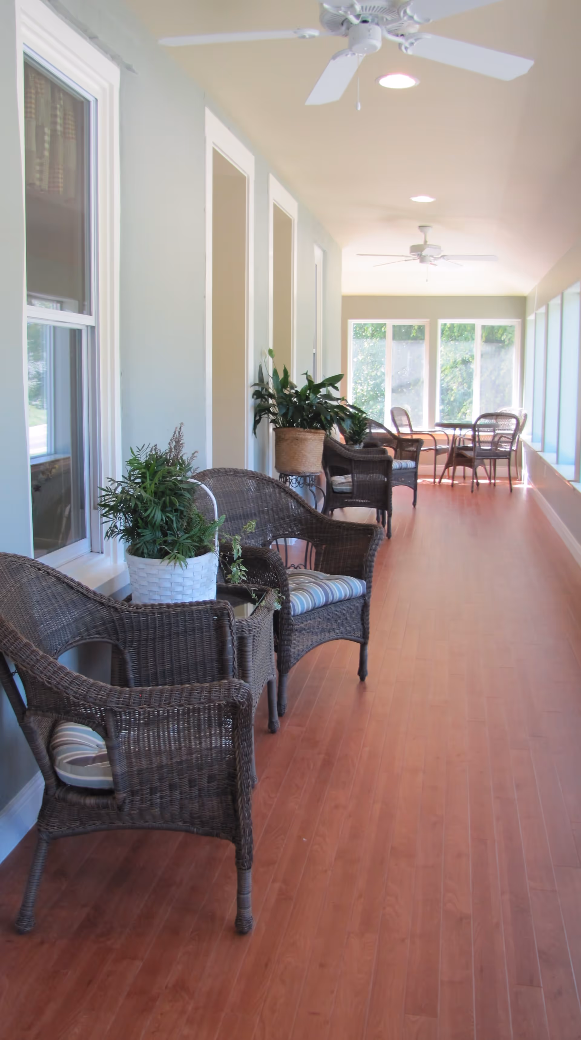 A bright, enclosed porch area with wooden flooring, wicker chairs with cushions, small tables, potted plants, ceiling fans, and large windows letting in natural light.