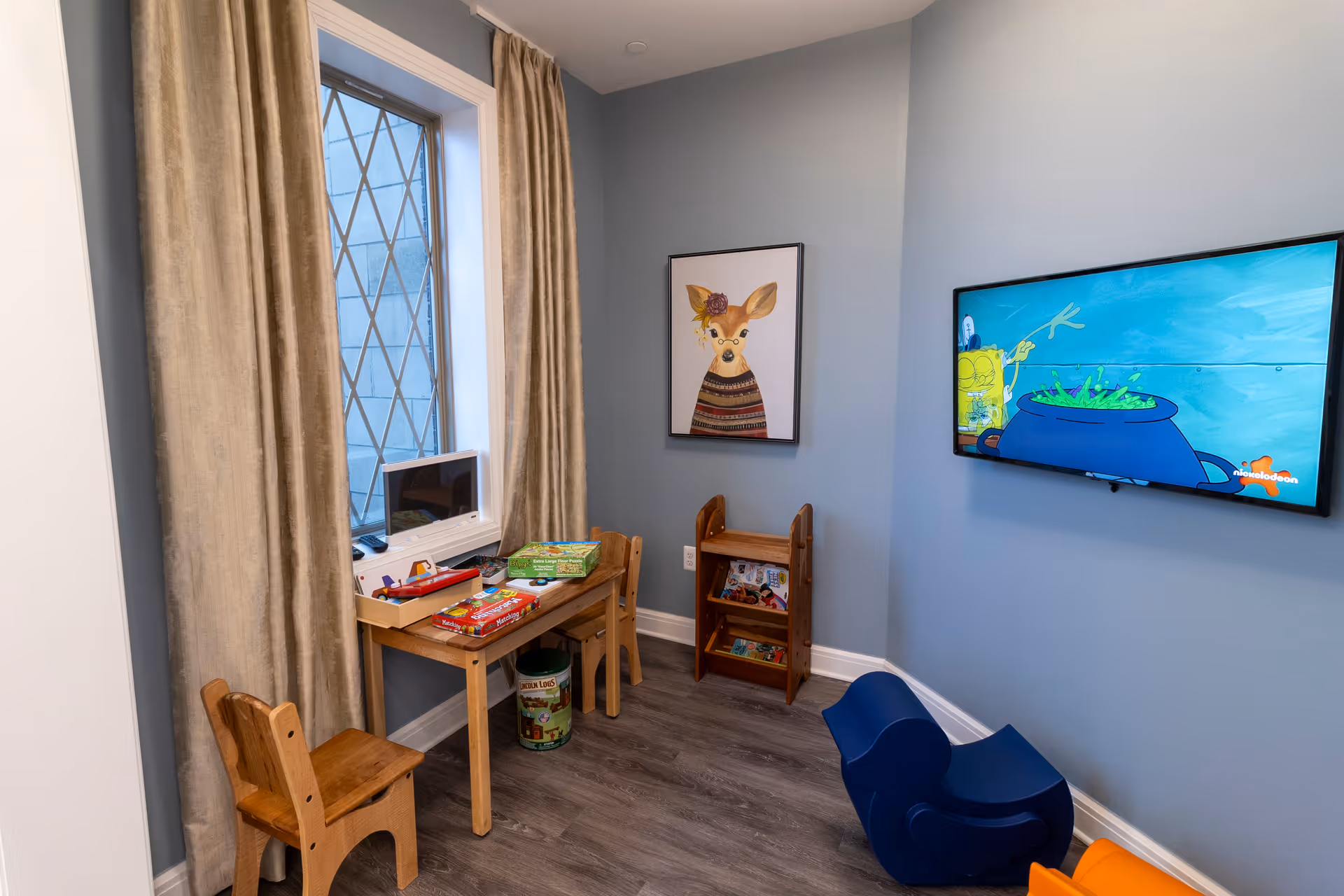 Small children's playroom with a wooden table and chairs, toys, a wall-mounted TV playing a cartoon, and framed animal artwork.