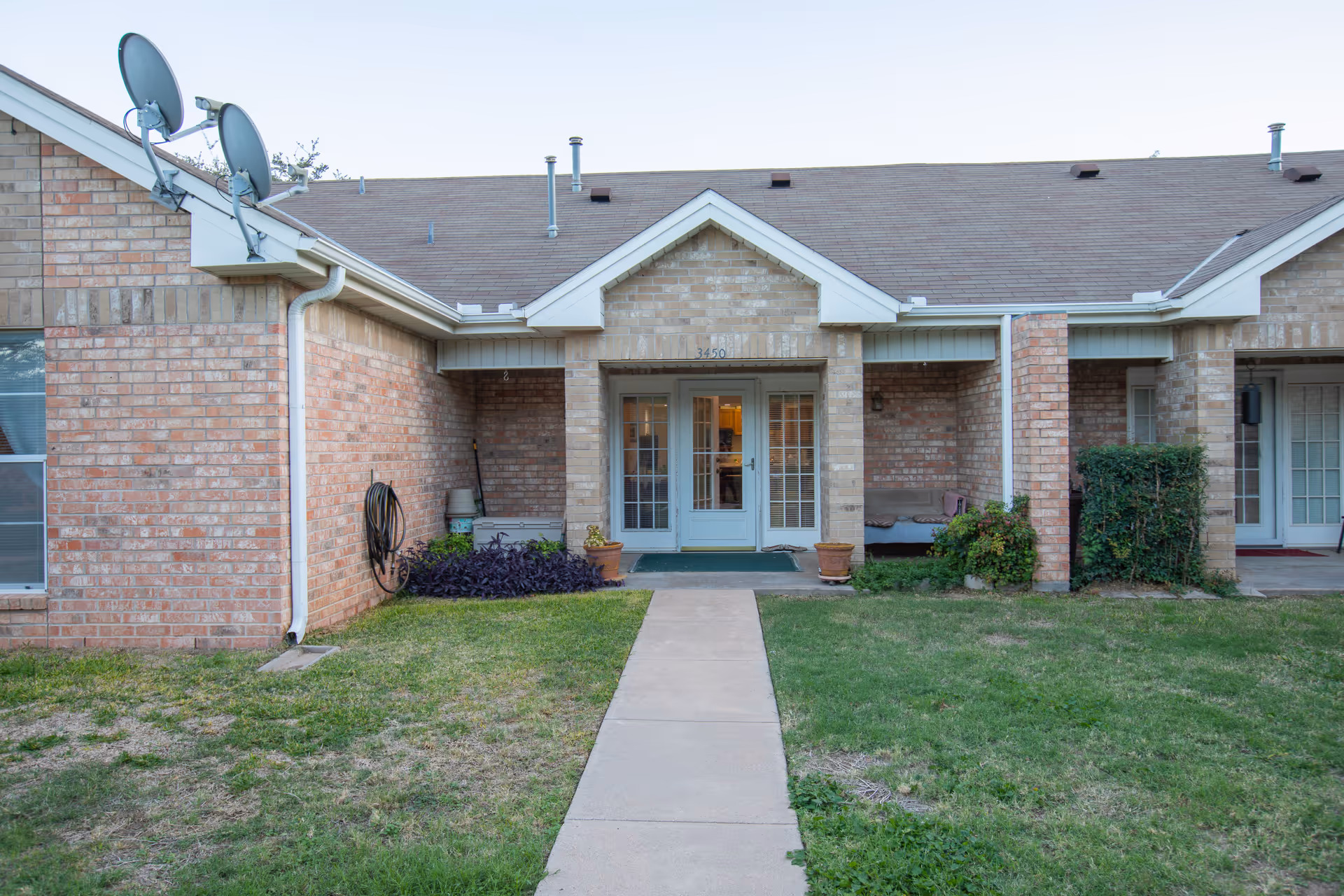 Brick single-story residential building entrance with a concrete walkway leading to glass double doors and small covered porches.