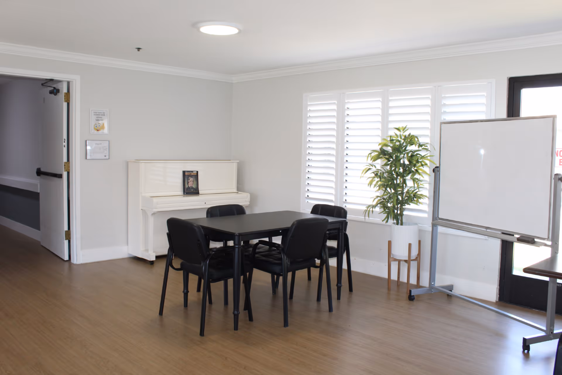 Bright common room with a black table and chairs, a white upright piano, a potted plant and a whiteboard by shuttered windows.