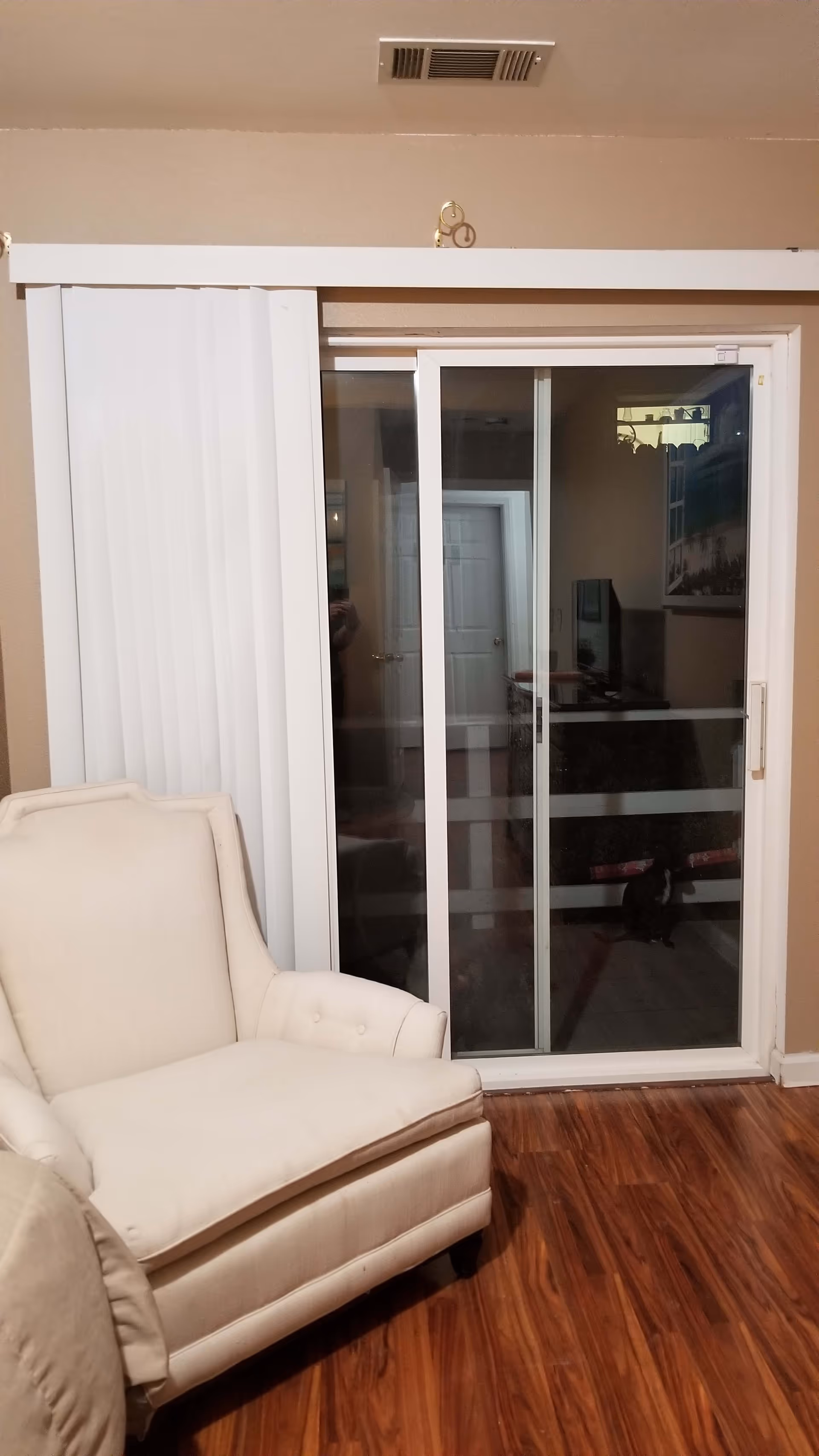 A beige upholstered armchair positioned on a wooden floor next to a sliding glass door with vertical blinds partially open. The door leads to an outdoor area with a white railing visible through the glass. The room has beige walls and a ceiling vent above the door.