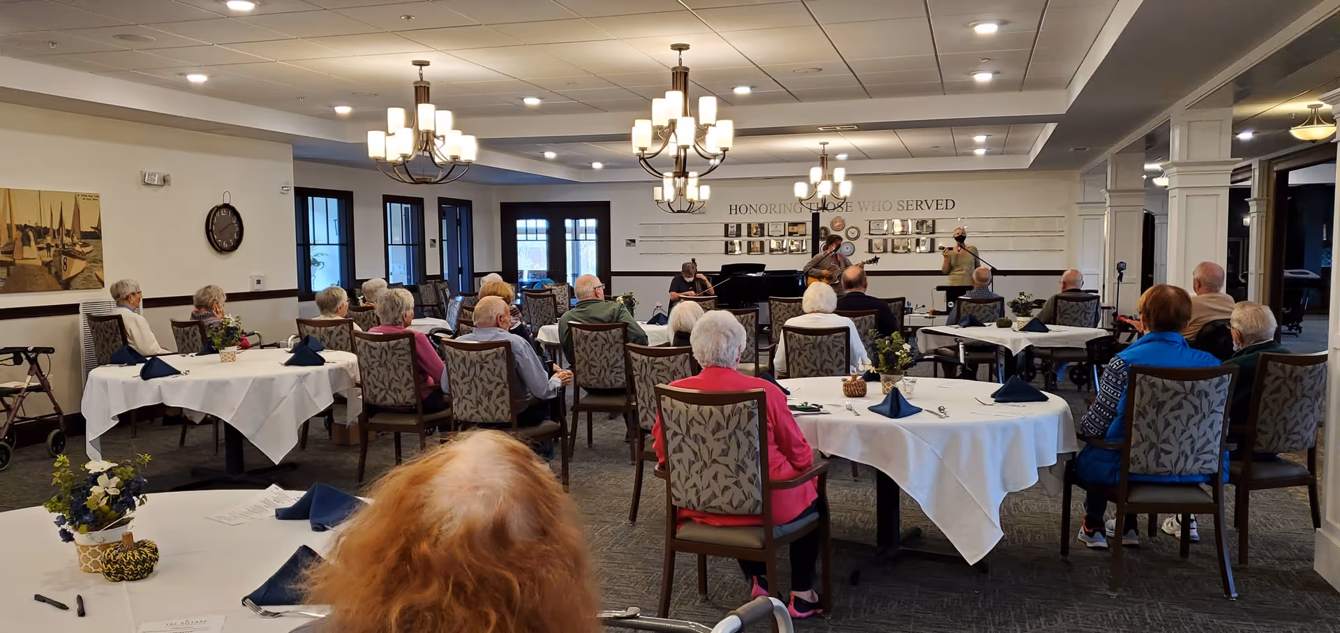 Seniors seated at round tables in a dining/activity room watching a small live musical performance at the front.
