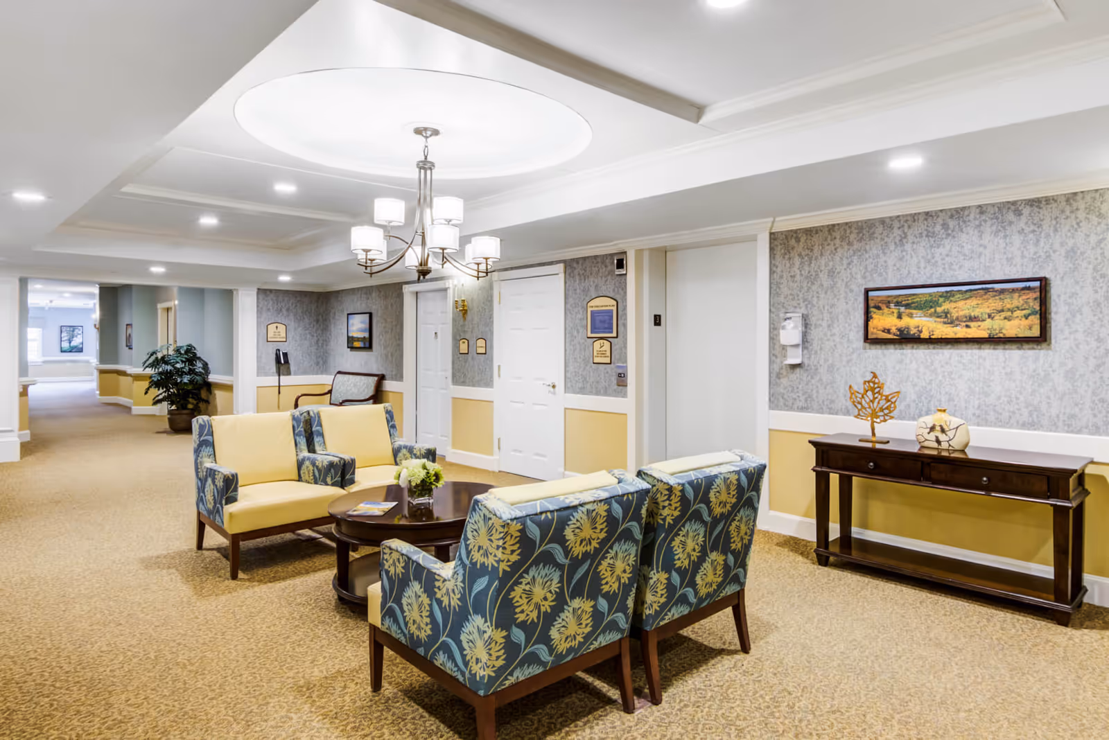 A well-lit senior living facility common area with patterned armchairs and sofas arranged around a round wooden coffee table. The walls are decorated with framed landscape paintings and a wooden console table with decorative items. The ceiling features recessed lighting and a chandelier.