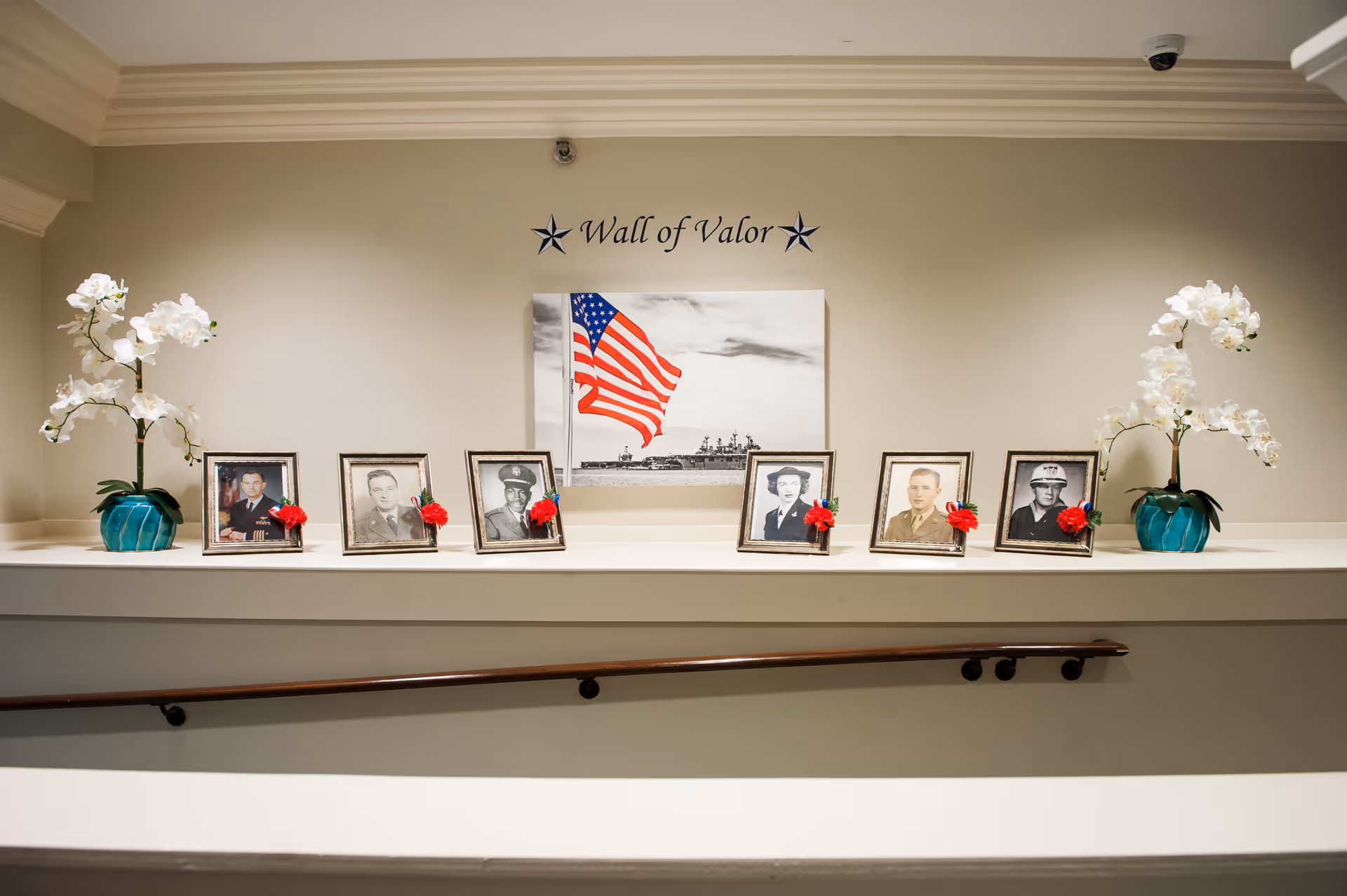 A memorial display titled 'Wall of Valor' featuring framed black and white photographs of military personnel with red carnations attached to each frame. The display includes a large picture of an American flag and a naval ship in the background, flanked by two white orchid plants in blue pots on either side.