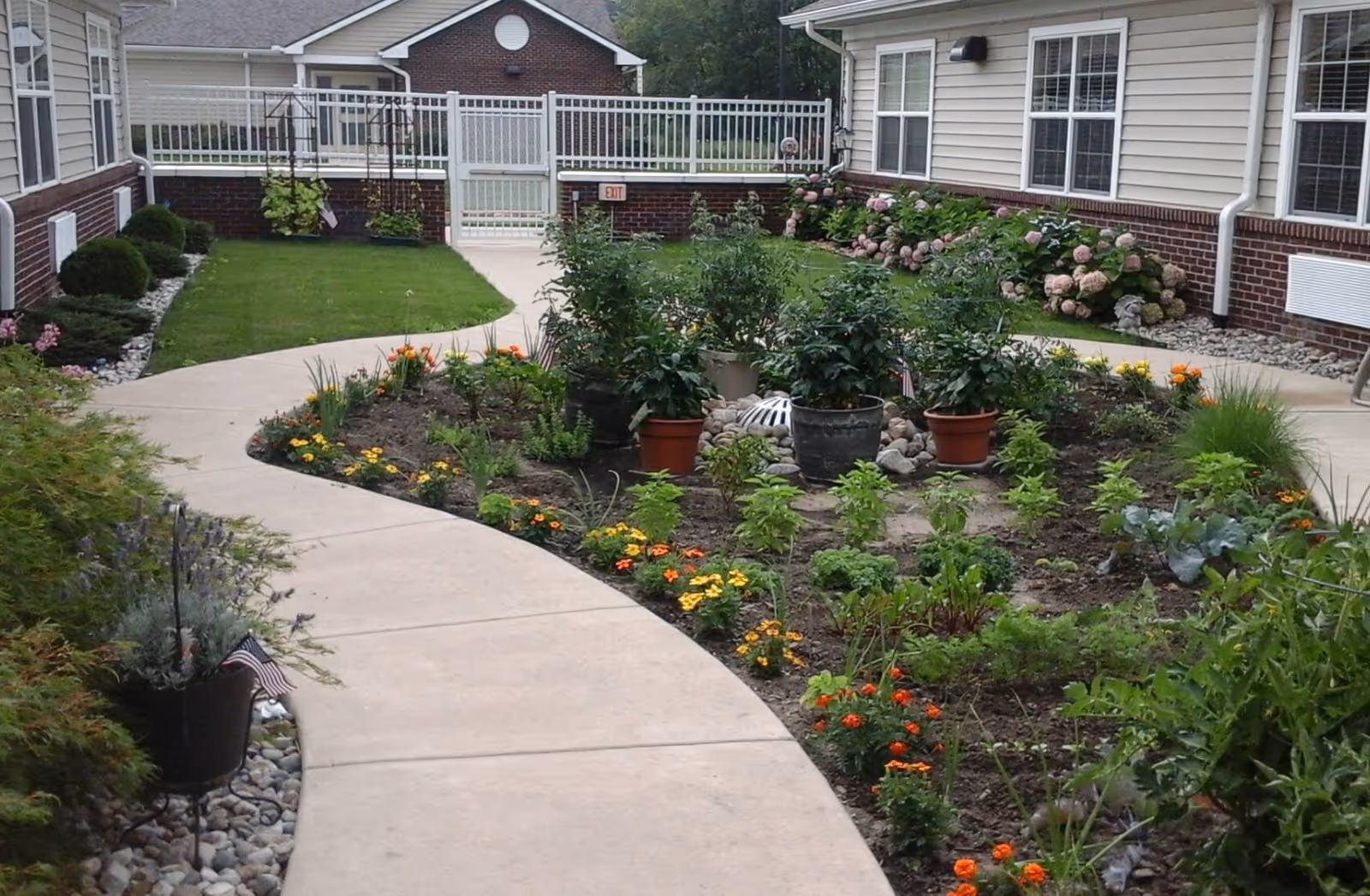 A landscaped outdoor garden area at The Willows at East Lansing featuring a curved concrete walkway surrounded by flower beds with various plants and flowers. The garden is bordered by buildings with windows and brick siding, and there is a white gate at the end of the walkway.