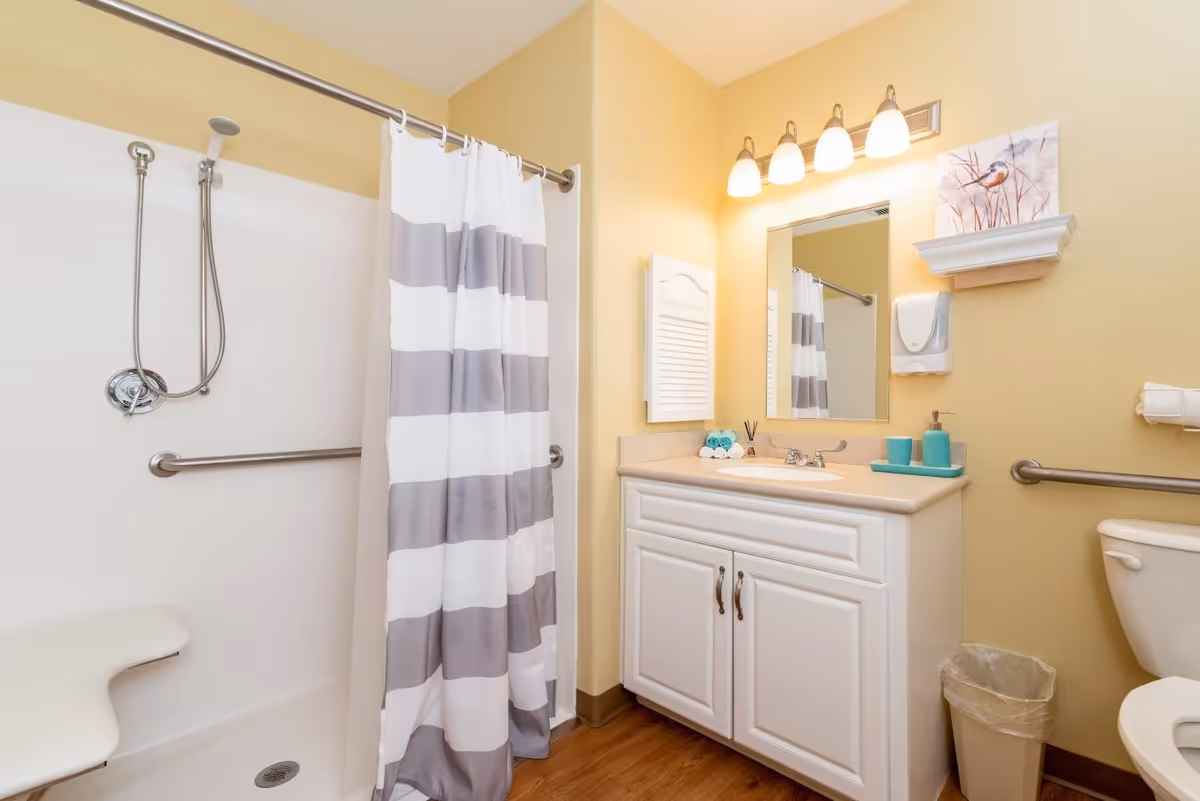 A clean bathroom with a white and gray striped shower curtain, a shower area with a handheld showerhead and grab bars, a white vanity with a sink, a mirror above the sink, a wall-mounted light fixture with four bulbs, a small shelf with a bird painting, and a toilet with a grab bar next to it. The walls are painted yellow and the floor has a wood-like finish.