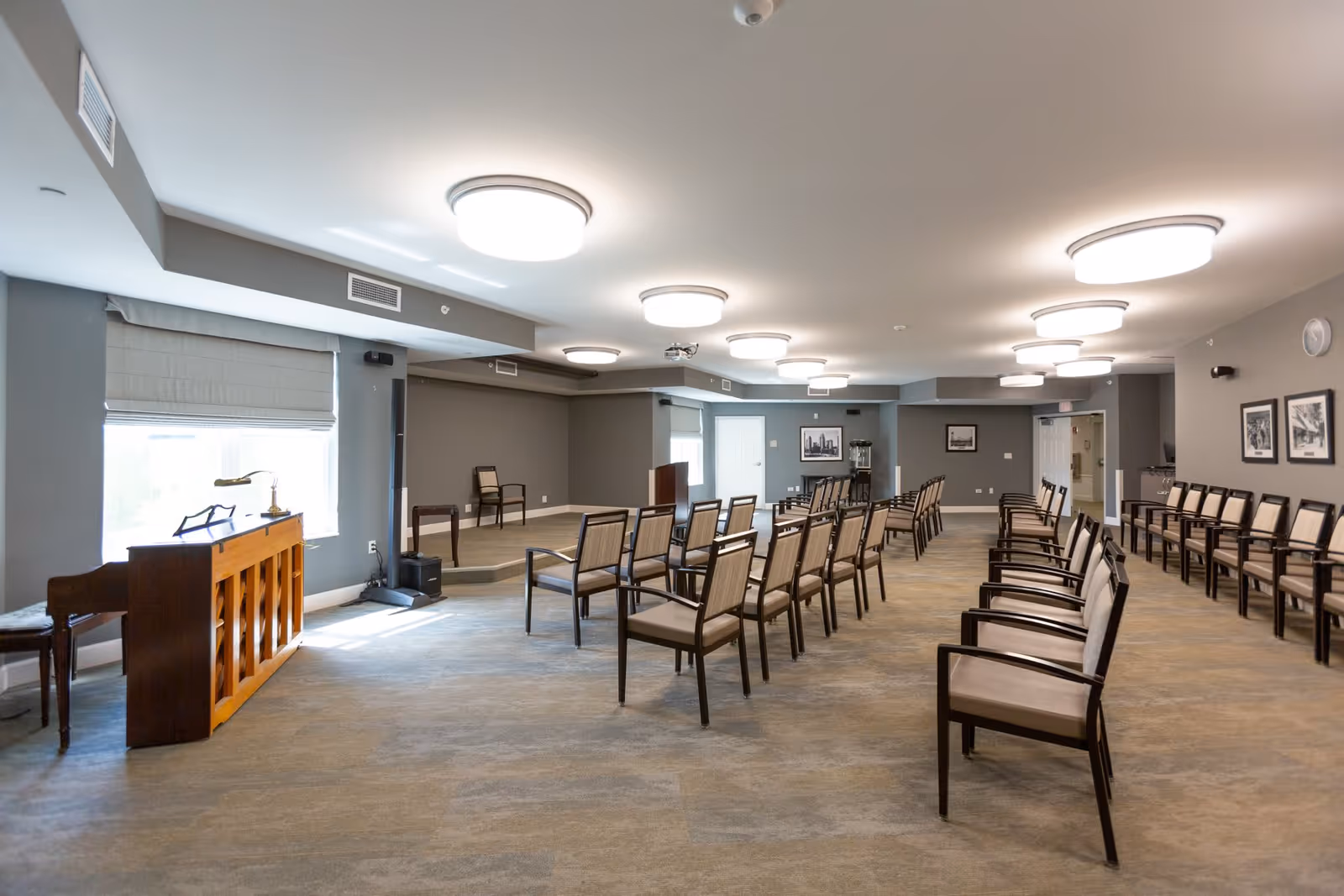 A spacious room with rows of chairs arranged facing a small stage area. The room has gray walls, carpeted floor, and several ceiling lights. There is a wooden piano on the left side near a window with a white blind, and framed pictures on the right wall. The room appears to be set up for a meeting or presentation.