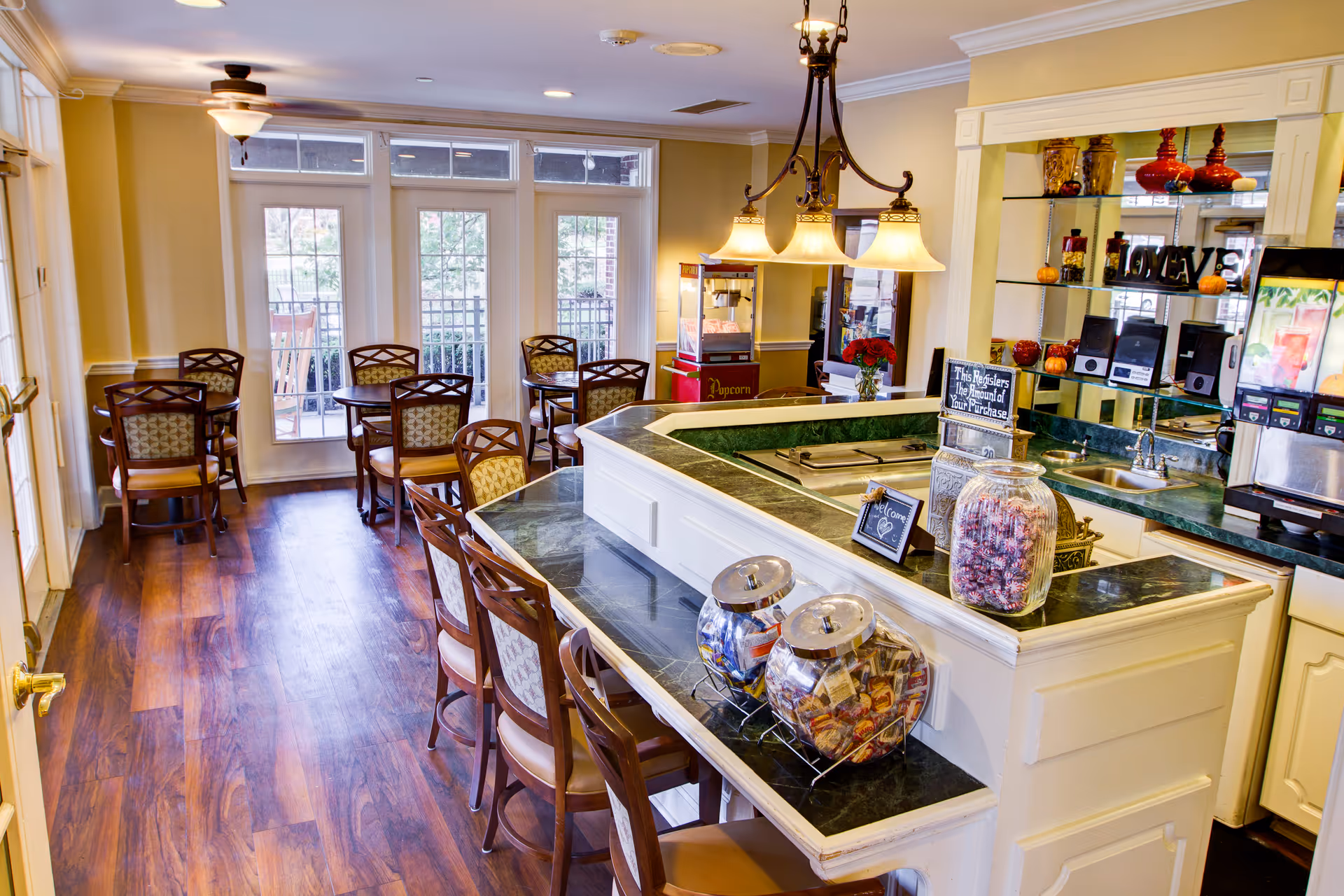 A bright and inviting dining area in a senior living facility with wooden floors, several round tables with chairs, a counter with bar stools, jars of snacks, a popcorn machine, and a beverage dispenser. The room has large windows and glass doors letting in natural light.