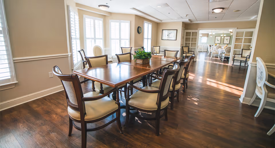 A bright dining room with a long wooden table surrounded by cushioned chairs. Large windows with white shutters allow natural light to fill the room. The floor is wooden, and there is a green plant centerpiece on the table. In the background, another dining area with similar furniture is visible through an open doorway.