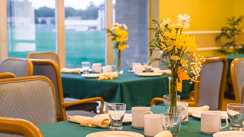 Dining room with tables covered in green tablecloths, set with white plates, cups, glasses, and yellow napkins. Each table has a vase with yellow flowers. Chairs with wooden frames and cushioned seats surround the tables. Large windows in the background let in natural light.