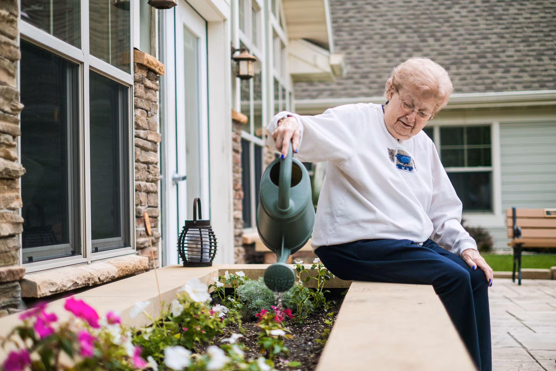 An elderly woman sitting on the edge of a raised garden bed outside a building, watering flowers with a green watering can. The building has stone and siding exterior walls, windows, and a door. There is a lantern on the ledge and a bench in the background on a paved patio area.