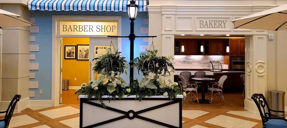 Indoor view of a senior living facility area with entrances to a barber shop and a bakery. The barber shop has a blue and white striped awning above the door, and the bakery has a white decorative frame around its entrance. In front of the entrances is a planter with green hanging and potted plants. There are chairs and tables visible inside the bakery area.
