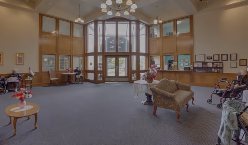 Spacious senior living facility common area with large windows and wooden paneling. The room features comfortable seating including armchairs and small tables with flower arrangements. Several elderly residents are seated, some with walkers. The entrance has tall glass doors letting in natural light. The walls display framed certificates and pictures.
