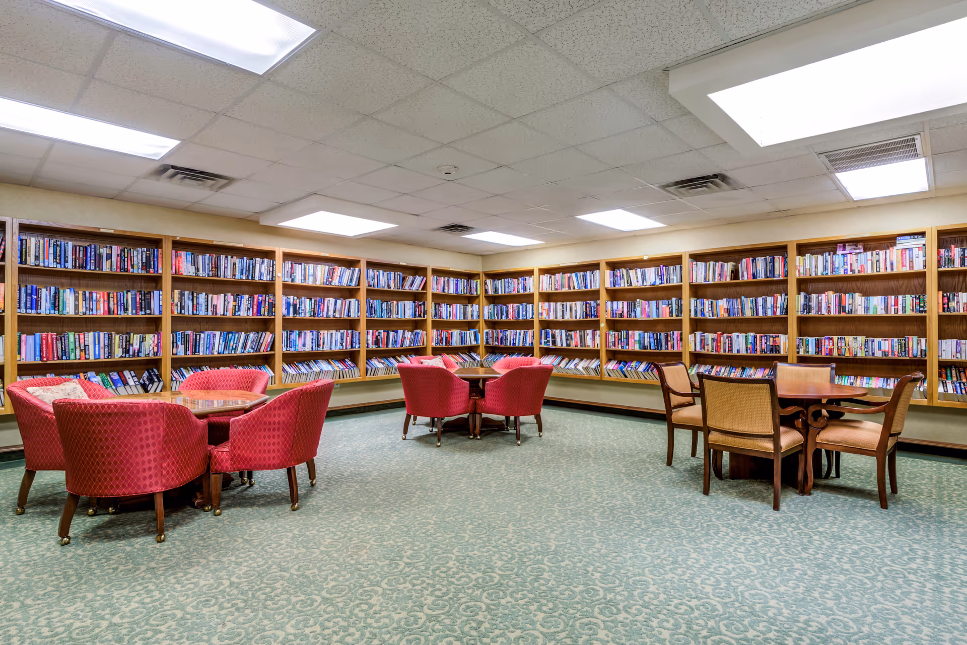 A spacious library room with green patterned carpet and walls lined with wooden bookshelves filled with books. There are several seating areas with round wooden tables surrounded by red upholstered chairs and beige wooden chairs. The ceiling has recessed lighting and a skylight.