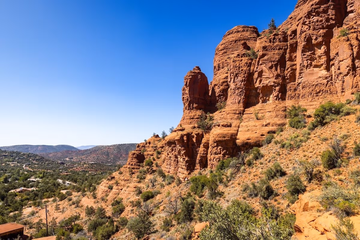 A scenic view of red rock formations and desert vegetation under a clear blue sky, with distant hills and scattered houses visible in the background.