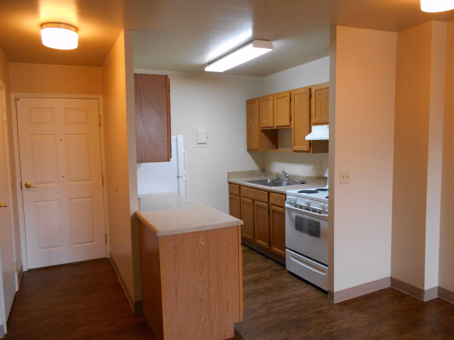 Interior view of a small kitchen area in a senior living facility with wooden cabinets, a white stove, a double sink, and a refrigerator. The kitchen is adjacent to a hallway with a closed white door and has wood flooring and beige walls.