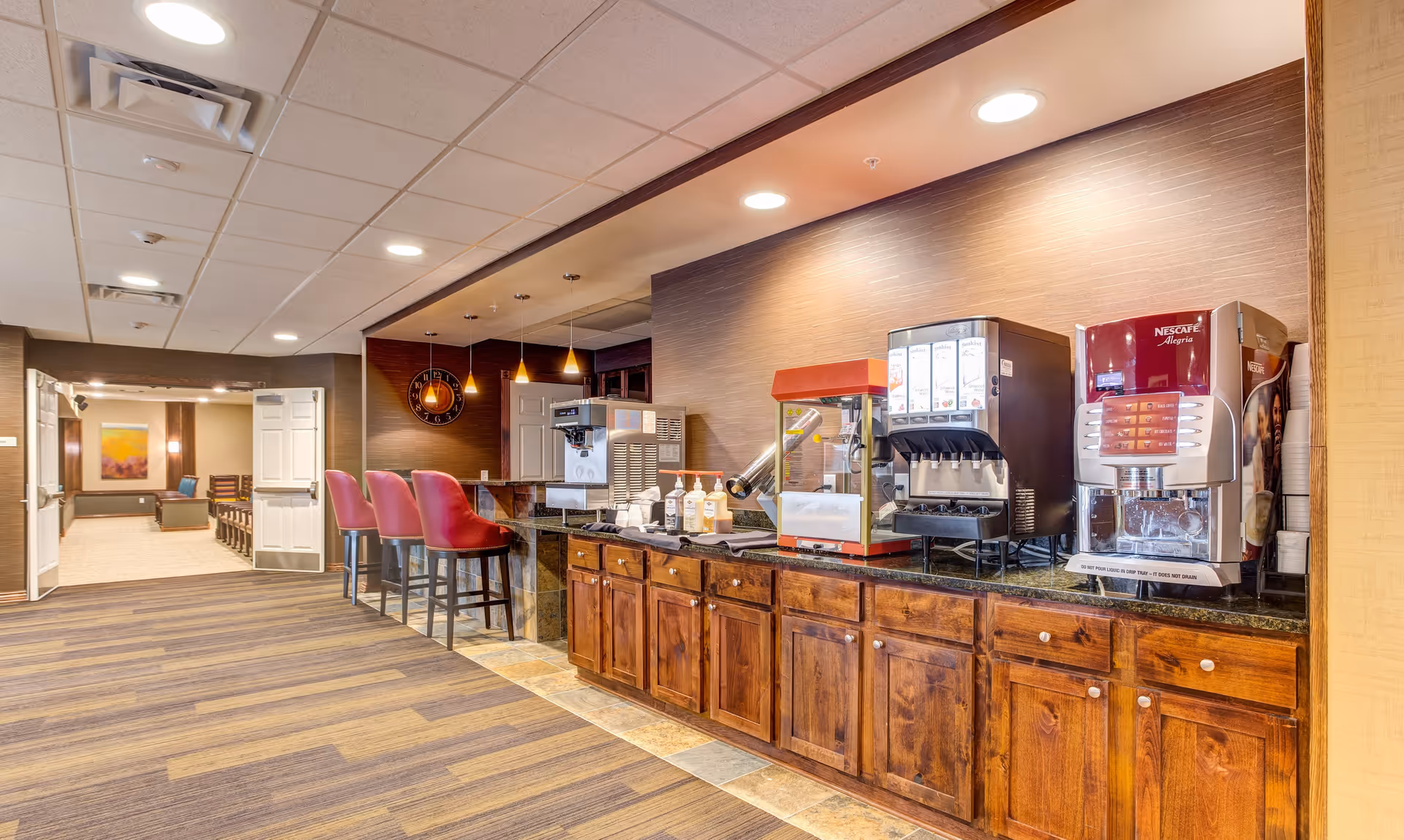 Interior view of a beverage station area in a senior living facility with coffee and drink machines on a wooden counter, red bar stools, and a hallway leading to a room with chairs and a painting on the wall.