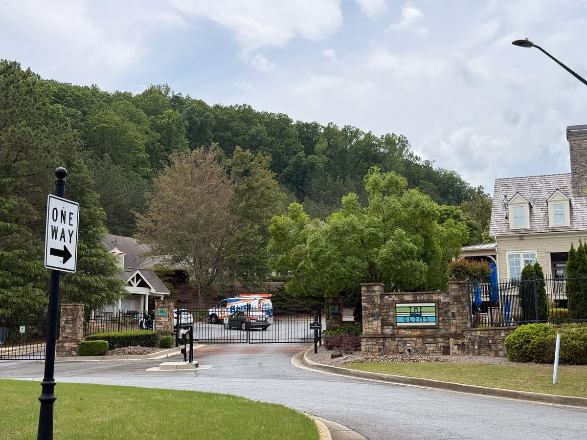 Gated entrance to a senior living community with a stone sign, driveway and buildings set against a tree-covered hillside.