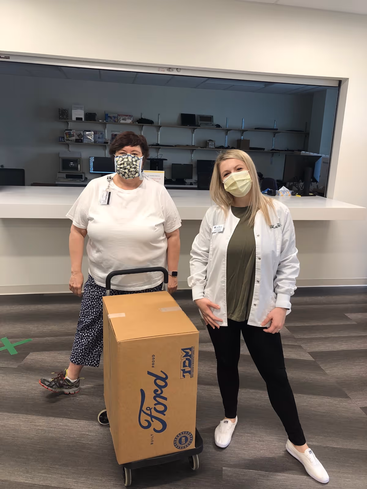 Two women wearing face masks standing indoors. One woman is pushing a dolly with a large cardboard box labeled 'Ford'. They are in a room with a counter and shelves with computer equipment in the background.
