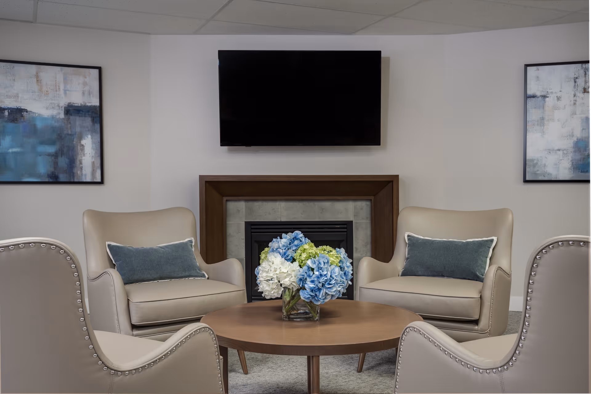 A cozy sitting area with four beige armchairs arranged around a round wooden coffee table holding a vase of blue, white, and green flowers. Behind the chairs is a fireplace with a wooden frame and a flat-screen TV mounted above it. Two abstract paintings with blue and gray tones hang on the white walls on either side of the fireplace.