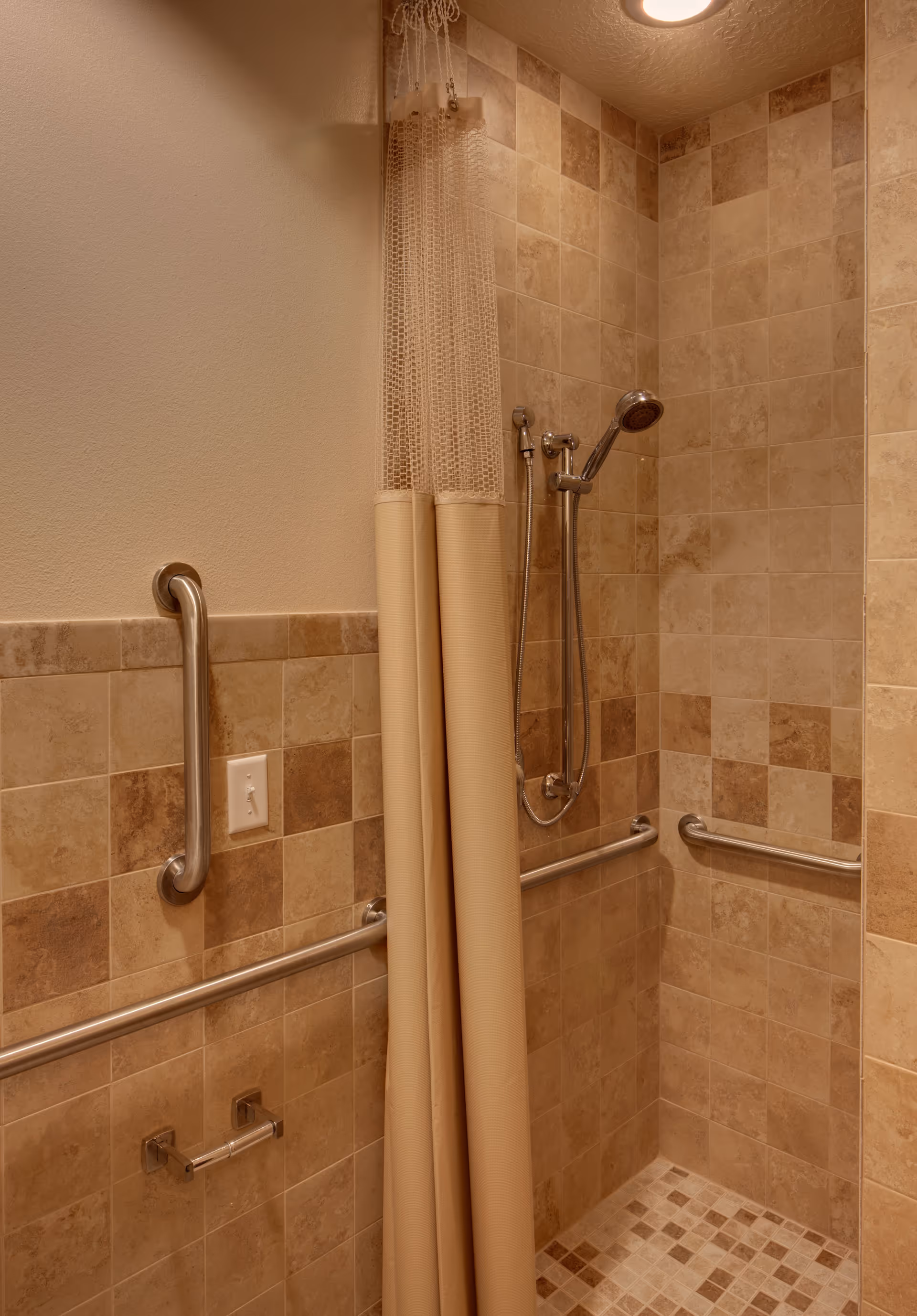 Tiled accessible shower with grab bars, a handheld showerhead, and a beige shower curtain.