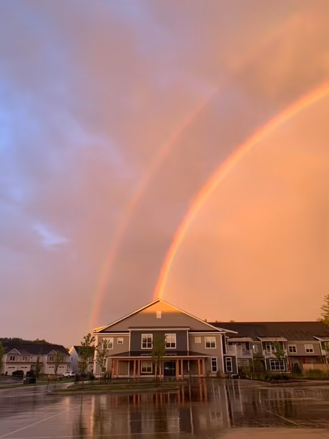 A residential building with a porch and multiple windows under a sky displaying a vivid double rainbow during sunset. The ground is wet, reflecting the building and the colorful sky. Other similar buildings are visible in the background.