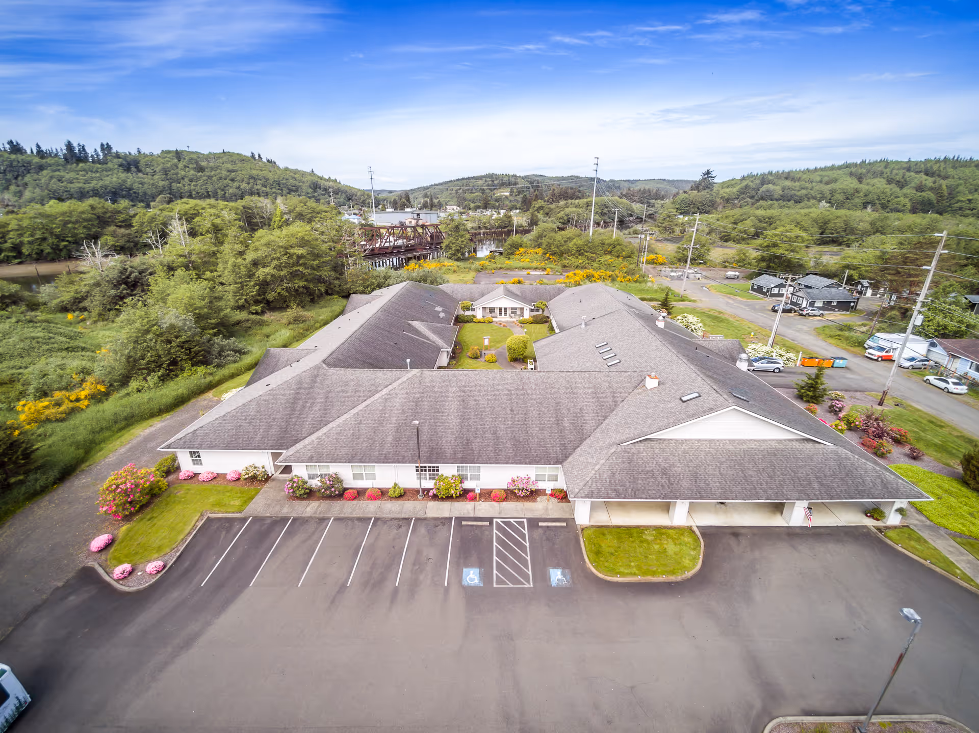 Aerial view of Riverside Place Memory Care, a single-story building with a parking lot, landscaped flower beds, and surrounding trees and hills.