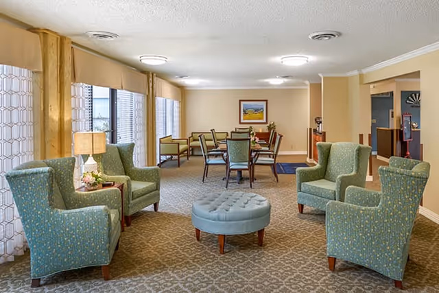 A spacious and well-lit common area with four green upholstered armchairs arranged around a round, tufted blue ottoman. In the background, there is a dining table with chairs and a framed landscape painting on the wall. Large windows with sheer curtains allow natural light to fill the room.