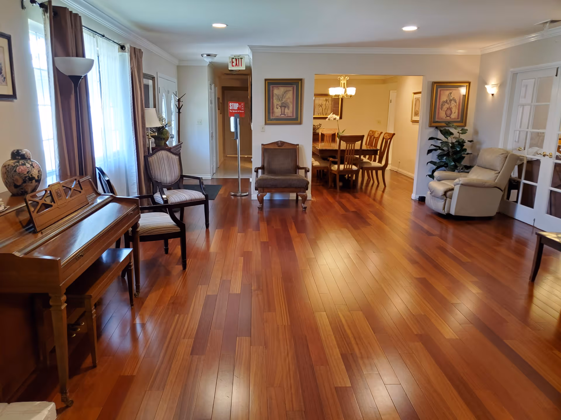 Spacious furnished common living area with hardwood floors, seating, a piano on the left, and a dining table visible in the background.