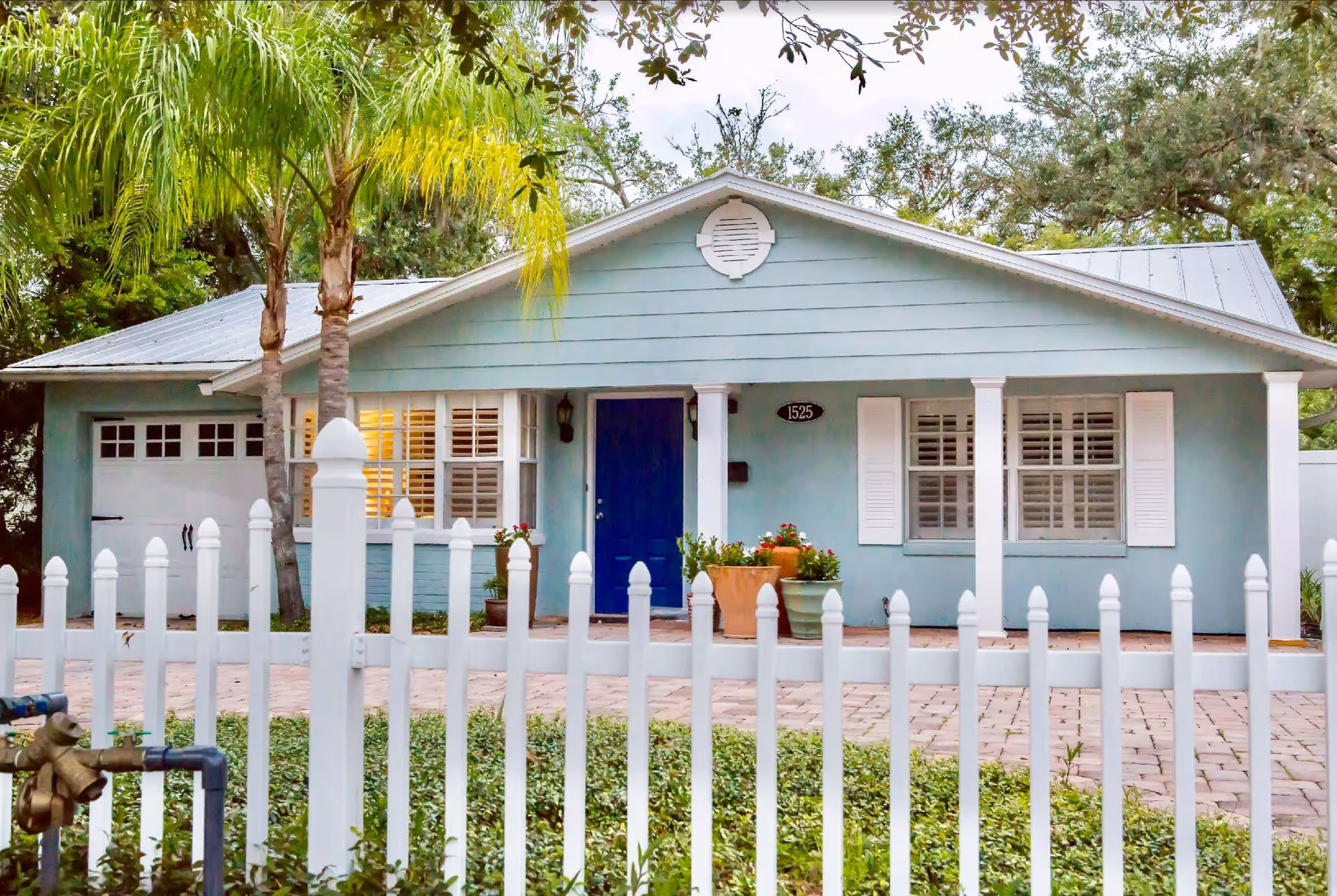 Front view of a light-blue single-story house with a blue door, white picket fence, garage, palm trees, and potted plants.
