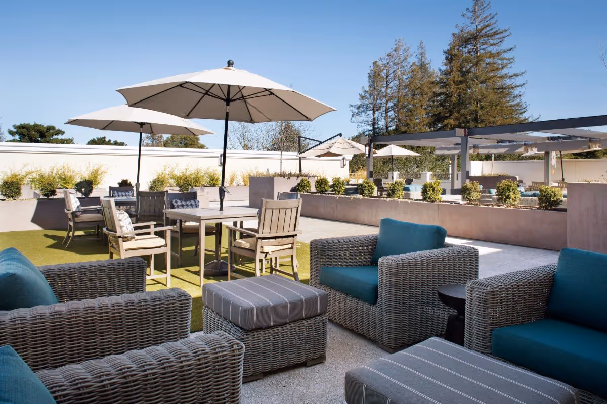 Outdoor patio area with wicker armchairs featuring teal cushions, matching ottomans, wooden tables and chairs, and large white umbrellas providing shade. The space is surrounded by planters with greenery and trees in the background under a clear blue sky.