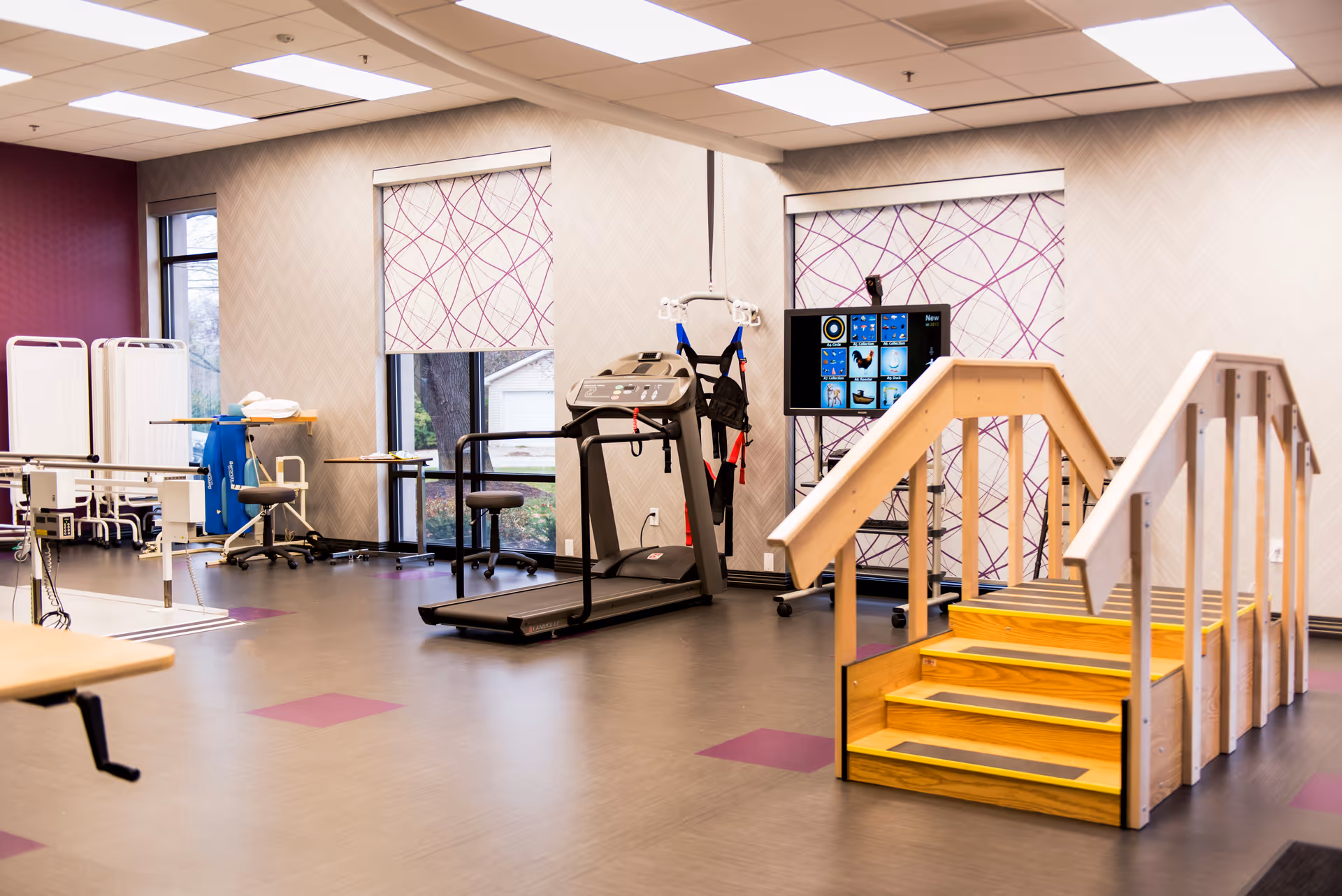 A rehabilitation or physical therapy room with exercise equipment including a treadmill, parallel bars, a wooden staircase for practice, and a screen displaying various images. The room has large windows with patterned blinds and a clean, spacious layout.