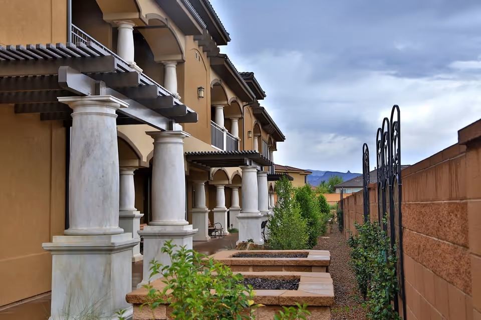 Outdoor view of The Retreat at Sunriver showing a walkway alongside a building with large white columns and pergolas. There are raised garden beds with plants and a brick wall with metal trellises on the right side under a cloudy sky.