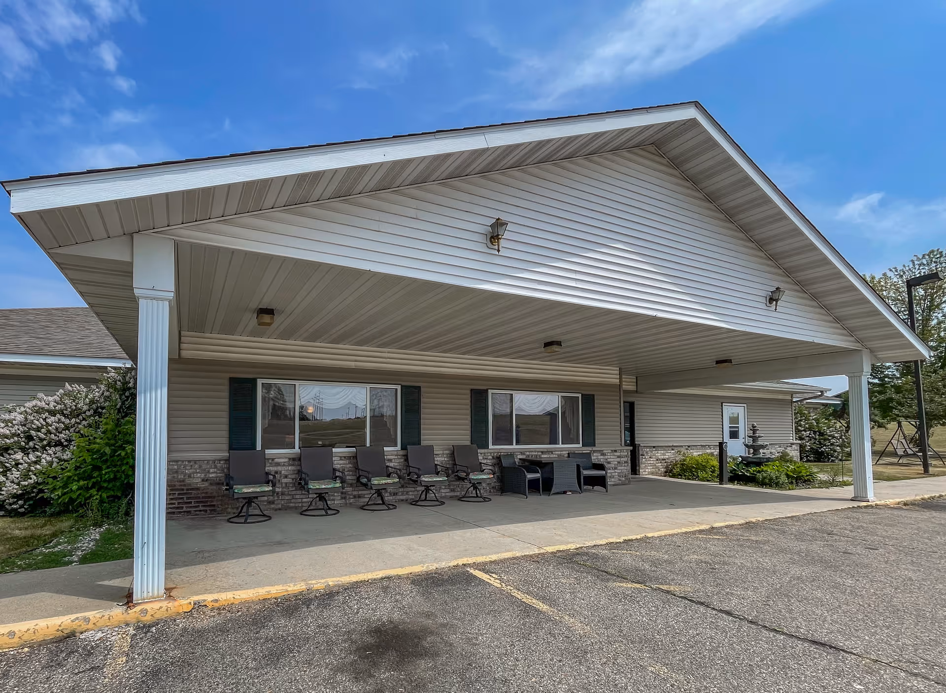 Covered entrance area of a single-story building with several chairs and a small table arranged on the concrete porch. The building has white siding, a sloped roof, and two large windows with dark shutters. There are some bushes and greenery around the building, and the sky is clear and blue.