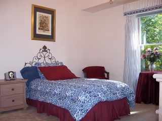 A bedroom with a bed covered in a blue and white patterned bedspread and red pillows. There is a wooden nightstand with a clock and a framed picture on the wall above the bed. A window with white curtains and a valance lets in natural light, and a round table with a red tablecloth and a vase of flowers is near the window.