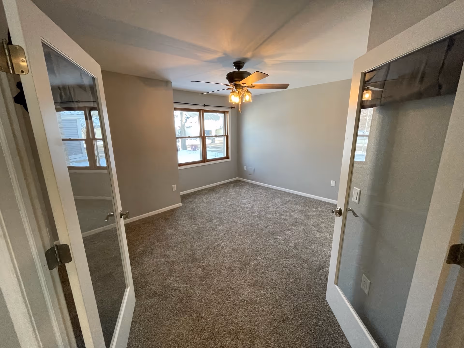 Empty room with beige carpet, light gray walls, a ceiling fan with lights, and a large window letting in natural light. The room is viewed through double glass-paneled doors.