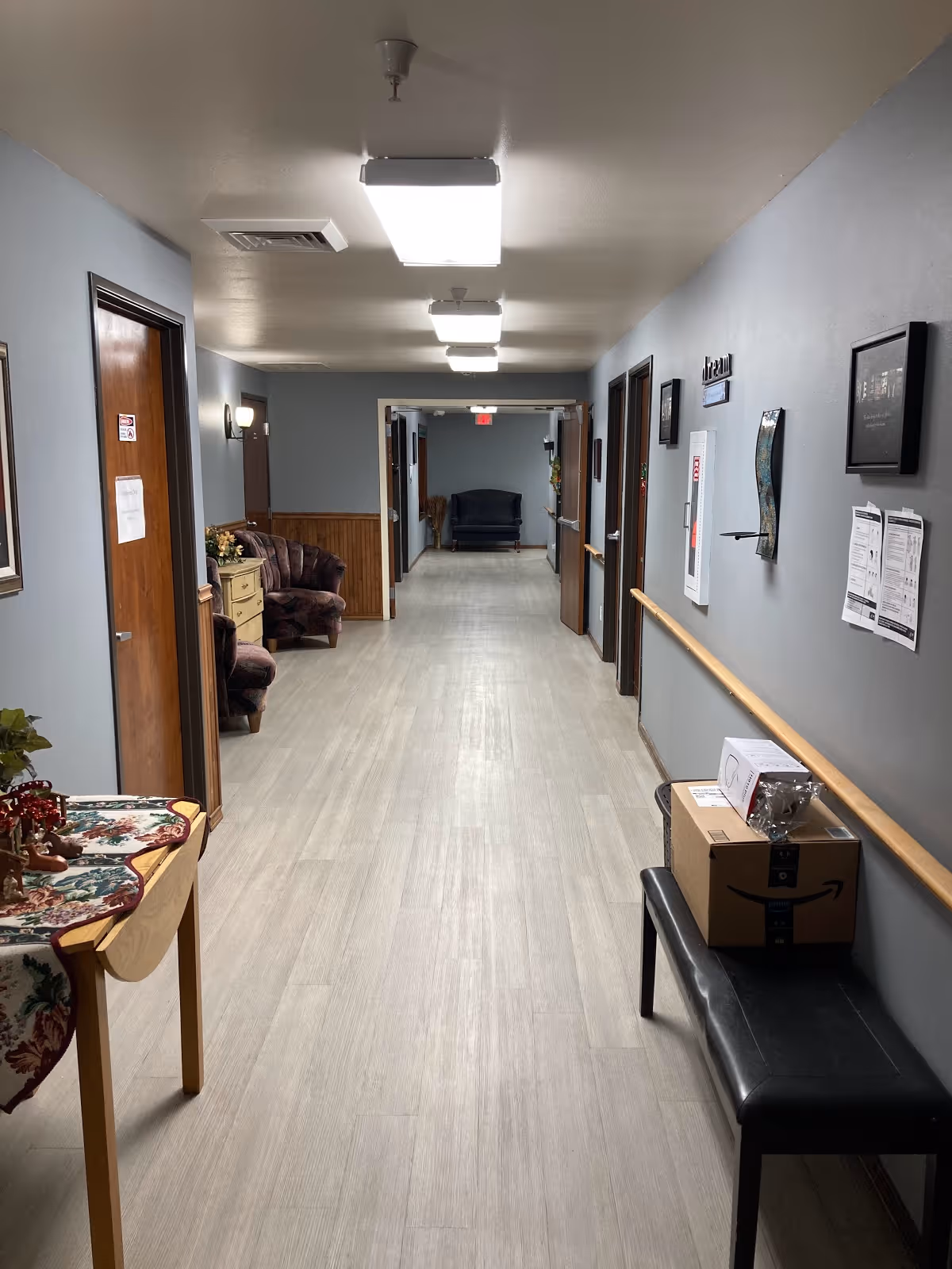 Long interior hallway with seating areas, room doors, a handrail, and packages on a bench.