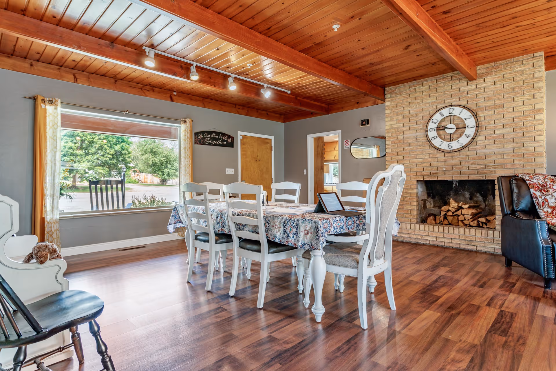 A cozy dining area with a wooden ceiling and hardwood floors. The room features a rectangular dining table covered with a floral tablecloth surrounded by six white chairs. There is a large window with yellow patterned curtains letting in natural light and showing greenery outside. A brick fireplace with stacked firewood and a large clock above it is on one wall. A black leather armchair with a patterned throw is partially visible near the fireplace. The walls are painted gray, and there are wooden doors and a mirror on the far wall.