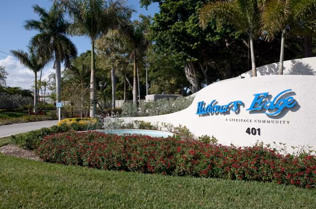 Entrance sign for Harbour's Edge, a Lifespace Community, with the address number 401. The sign is white with blue lettering and is surrounded by red flowering bushes and palm trees under a clear sky.