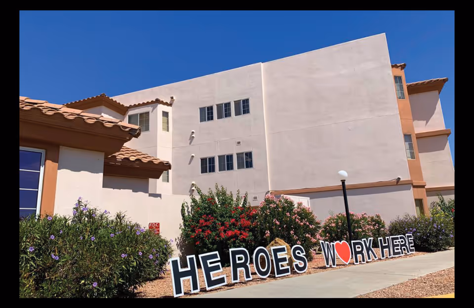 Exterior of a pale stucco multi-story retirement home with landscaping and a large sign reading 'HEROES WORK HERE'.