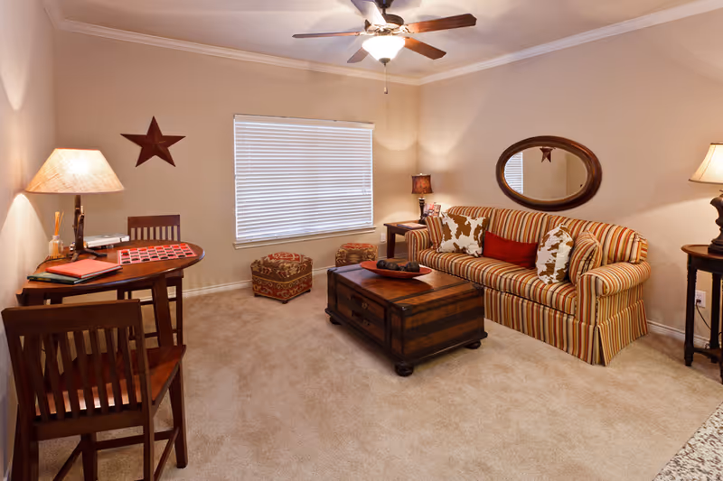 A cozy living room with beige walls and carpet, featuring a striped sofa with decorative pillows, a wooden coffee table, two patterned ottomans near a window with closed blinds, a small wooden table with a lamp and books, and a ceiling fan with a light fixture.