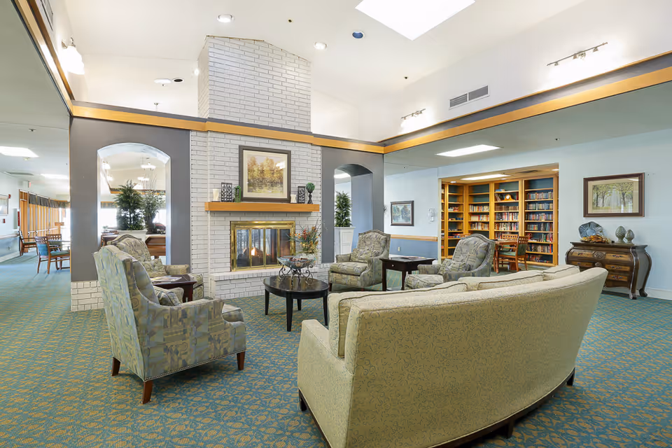A spacious senior living common room with sofas and armchairs arranged around a brick fireplace and a bookcase in the background.
