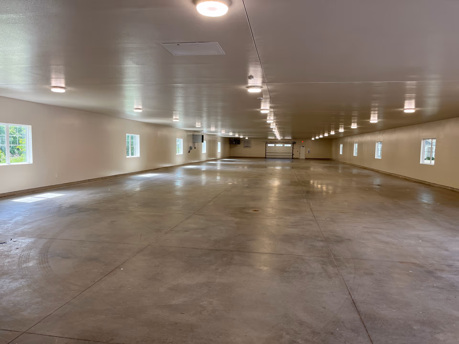 A large empty indoor space with polished concrete floors, white walls, and multiple evenly spaced windows along both sides. The ceiling has numerous round recessed lights and ventilation panels. At the far end, there is a garage-style door and a regular door beside it.