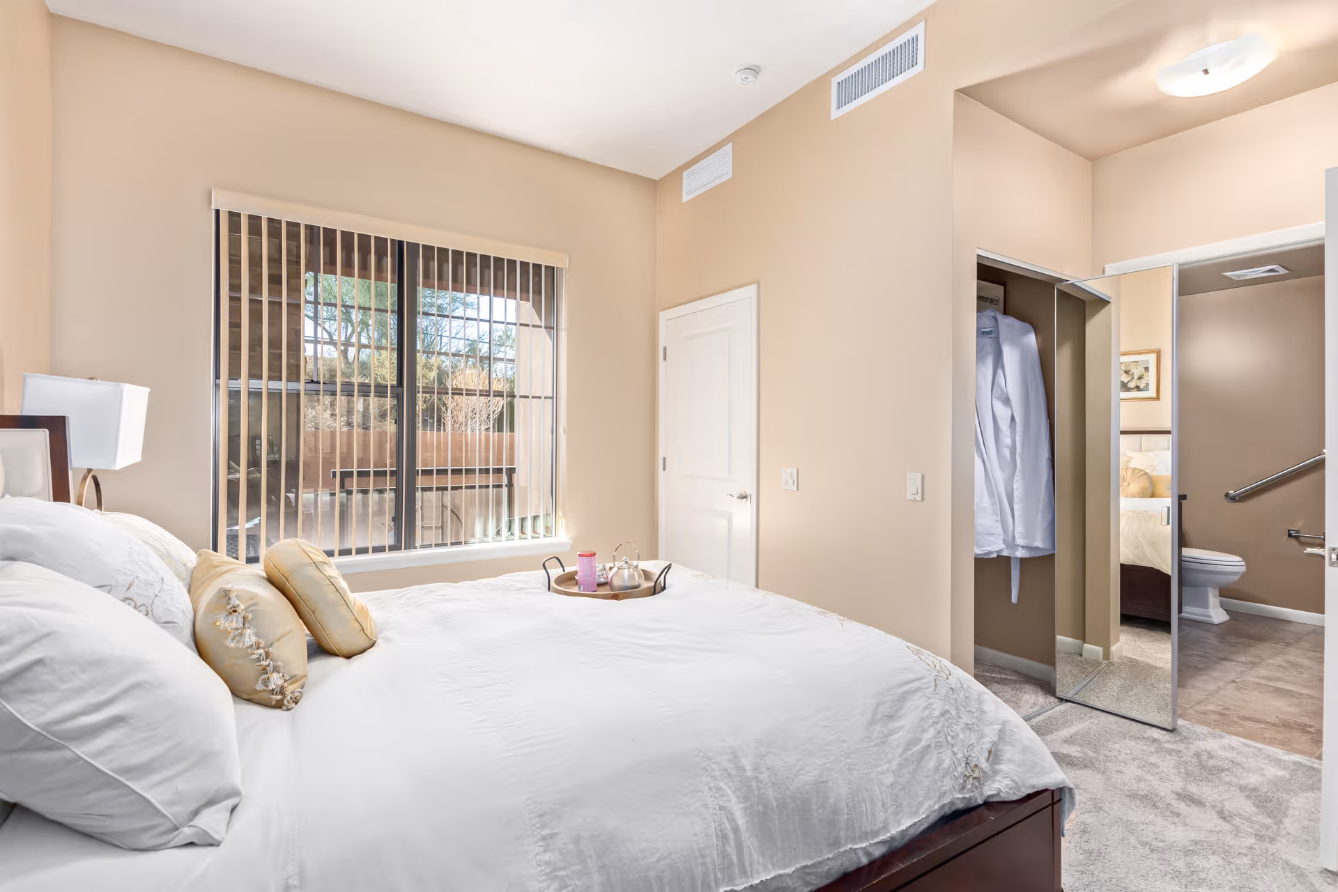 Light-filled bedroom with a made bed and decorative pillows, a large window with vertical blinds, an open closet with mirrored door, and a view into the bathroom.