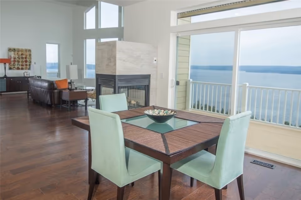 A bright living and dining area with a square wooden dining table with four light green upholstered chairs. The table has a decorative bowl in the center. Behind the table is a modern double-sided fireplace with a light stone surround. The room has large windows and a sliding glass door that opens to a balcony with a view of a large body of water. The living area has dark wooden floors, a brown leather sofa, side tables with lamps, and a decorative wall piece.